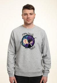 Muž oblečený v šedé mikině s grafikou dvou žen zády k sobě uvnitř kruhového rámu s designem větví a pavučiny.