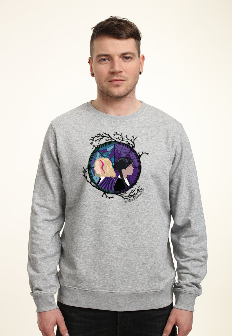 Muž oblečený v šedé mikině s grafikou dvou žen zády k sobě uvnitř kruhového rámu s designem větví a pavučiny.