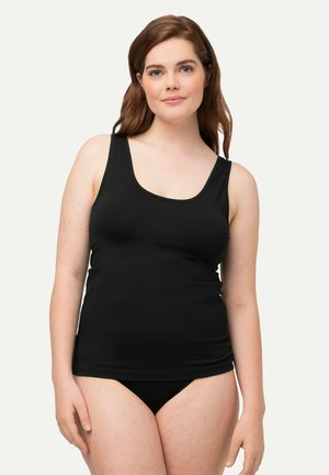 Donna dalla pelle chiara e capelli castani indossa una canottiera nera e biancheria intima nera, in piedi contro uno sfondo chiaro e semplice.