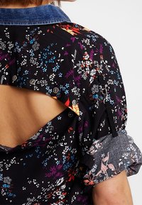 Chemise noire à motifs floraux avec col en denim, dos ouvert avec boutons-pression et manches retroussables maintenues par une lanière boutonnée.