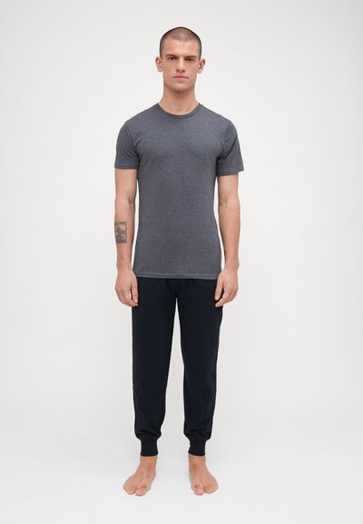 Graues kurzärmliges T-Shirt in schlanker Passform, kombiniert mit schwarzen Jogginghosen mit Bündchen an den Knöcheln. Das Modell steht barfuß und aufrecht.