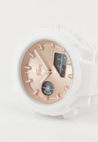 Montre numérique blanche avec un cadran rose doré, de forme circulaire, et des marqueurs d'heure blancs. Ses caractéristiques incluent un affichage numérique et un indicateur "Soleil".