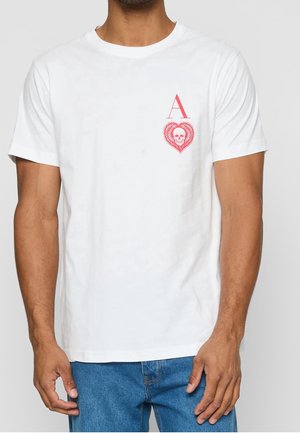 Mężczyzna ubrany w biały T-shirt z czerwonym wzorem Asa kier, przedstawiającym czaszkę w sercu, w połączeniu z niebieskimi dżinsami.