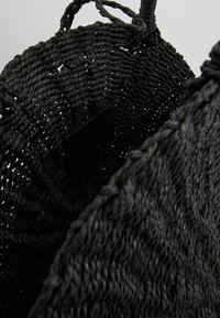 Svart vävd väska med en texturerad design, med en öppen topp och två robusta handtag. Innehåller en svart etikett inuti.