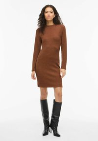 Vestito marrone in maglia con maniche lunghe e scollo rotondo, caratterizzato da una silhouette aderente. Indossato con stivali alti in pelle nera.