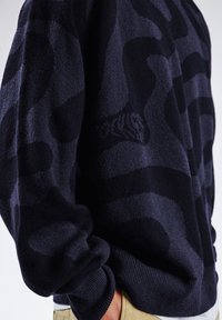 Laivastonsininen neulepaita, jossa on teksturoitu kangas, rohkea musta maastokuosi, resorireunat hihansuissa ja helmassa sekä hillitty logoyksityiskohta edessä.