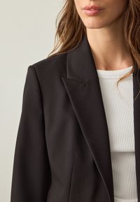 Černé sáčko s výraznými klopami, na míru střižené a s hladkou texturou, doplněné bílým žebrovaným topem pod ním.