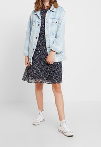 Hellblaue Jeansjacke über einem navy-blauen blumigen Kleid mit schwingendem Rock, kombiniert mit weißen High-Top-Sneakern.