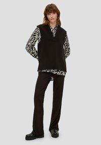 Top senza maniche nero con scollo a V sopra una camicia a righe bianco e nera, abbinato a pantaloni neri svasati e scarpe nere spesse.