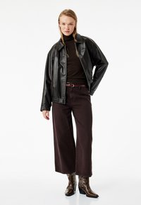 Schwarze Lederjacke über einem braunen Rollkragenpullover, kombiniert mit weit geschnittenen bordeauxroten Hosen und braunen bestickten Cowboy-Stiefeln.