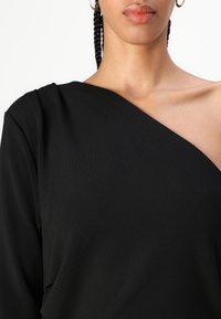 Svart asymmetrisk topp med en myk tekstur, som har en lang erm og en eksponert skulderdesign. Glatt overflate og avslappet passform.
