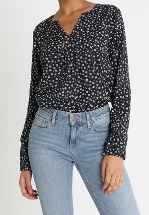 Kvinde iført en sort bluse med lange ærmer med hvide abstrakte pletter og lyseblå højttaljede jeans, stående mod en ensfarvet baggrund.