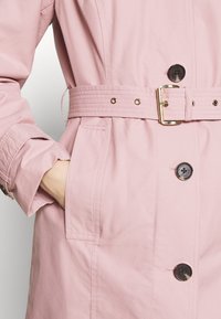 Trench-coat rose clair avec une taille ceinturée, des boutons noirs et une main partiellement glissée dans une poche latérale.