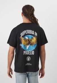 Črna bombažna majica s pisanimi grafičnimi motivi orla, besedilom "SUPERBIA IN PROELIO" in logotipom na spodnjem središču hrbta.
