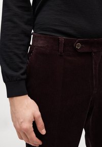 Burgundy kordbársony nadrág, függőleges bordákkal, fém gombos záródással és oldalsó zsebekkel, fekete, hosszú ujjú inggel párosítva.