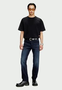 Homem vestido com uma t-shirt preta, jeans azul escuro, cinto preto, botas pretas, óculos de sol de tonalidade laranja e relógio de pulso prateado, de pé contra um fundo branco.