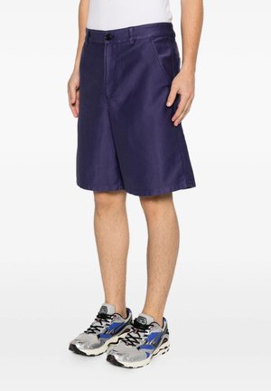 Uomo con pantaloncini blu navy e scarpe da corsa grigio-blu in piedi su sfondo bianco.