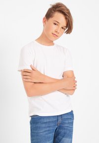 T-shirt bianco a maniche corte abbinato a jeans denim blu, con una vestibilità classica e una texture morbida. Il modello è in posa con le braccia incrociate.