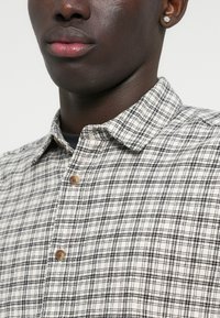 Švarkas su mygtukais, pagamintas iš tekstūruotos medžiagos, su smėlio ir juodos spalvos languotu raštu, turintis apykaklę ir medinius mygtukus.