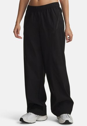 Person wearing loose black pants with elastic waistband and white athletic shoes standing on a plain background.