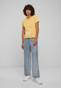 Stribet gul og hvid kortærmet t-shirt parret med lyseblå vide jeans og hvide sneakers, sat mod en lys grå baggrund.