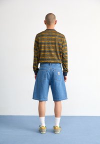 Long-sleeve green and orange striped shirt, light blue denim shorts, white socks, and multicolor sneakers. Plain background.