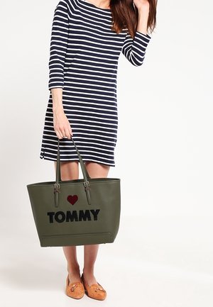 Olivgrüne Tragetasche mit schwarzem "TOMMY"-Text und rotem Herz, mit doppelten Henkeln und einer glatten, strukturierten Oberfläche. Gestreiftes Kleid zu sehen.