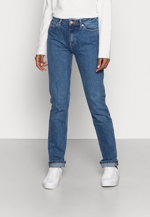 Person, die hochgekrempelte blaue Jeans, ein weißes Langarmshirt und weiße Sneaker trägt und vor einem einfarbigen hellen Hintergrund steht.