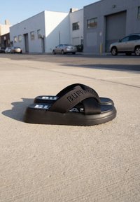 Sandales compensées noires à enfiler avec des brides croisées et le logo "BUFFALO", posées sur un trottoir en béton à proximité d'une rue industrielle.