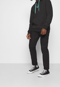 Felpa nera con cordini turchesi, pantaloni neri e scarpe da ginnastica nere. La felpa presenta un piccolo logo di un colore turchese abbinato.