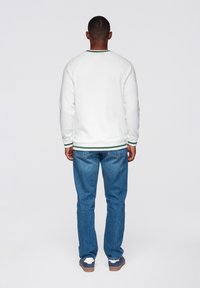 Vit sweatshirt med raglanärmar och grön rand vid kragen, kombinerad med blå denimjeans och bruna sneakers. Bakifrån.