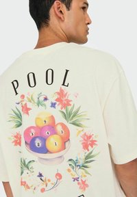 Camiseta de color crema con un gráfico colorido de bolas de billar en un cuenco, rodeadas de acentos florales, con el texto "POOL" en la parte superior.