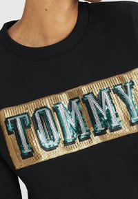 Czarna bluza z dużym haftowanym cekinami wzorem "TOMMY" w kolorach złotym i zielonym, z teksturowanym, prążkowanym dekoltem.