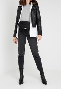 Veste de moto en cuir noir portée sur un t-shirt gris, associée à un jean noir taille haute et des bottines noires. Comprend un sac-banane noir avec logo.
