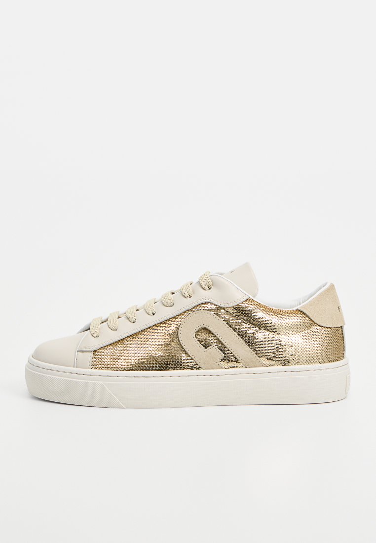 Zapatillas beige con un panel lateral texturizado en metal dorado, cordones en beige claro y puntera lisa. Suela blanca gruesa y forma redondeada.
