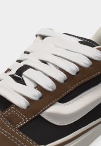 Κοντινό πλάνο ενός αθλητικού παπουτσιού με λευκά κορδόνια, καφέ σουέτ λεπτομέρειες και λευκή δερμάτινη ρίγα στο πλάι σε μαύρη βάση.