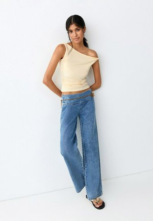 Žena u bež topu s jednim ramenom, širokim plavim trapericama s remenom od užeta i crnim japankama, naslonjena na bijeli zid.