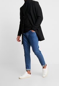 Cappotto nero su un dolcevita nero, abbinato a jeans in denim azzurro chiaro e sneakers bianche con dettagli bordeaux. Illuminazione piatta, sfondo neutro.