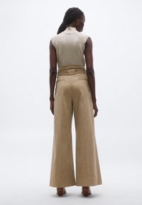 Pantaloni largi bej cu talie înaltă, având un detaliu cu curea și buzunare laterale. Țesătura pare netedă, cu finisaj mat.