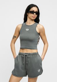 Γυναίκα που φοράει γκρι αμάνικο crop top και αντίστοιχα σορτς με κορδόνι, σκούρα γυαλιά ηλίου, με τατουάζ στο πάνω μέρος του αριστερού μπράτσου, τα χέρια στις τσέπες.