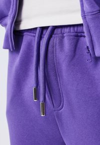 Primer plano de pantalones de chándal morados con cordones y puntas de metal, usados con una camiseta blanca y una chaqueta morada con cremallera a juego.
