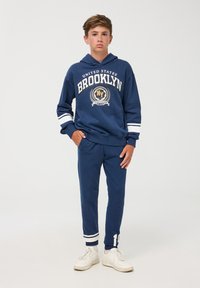 Granatowy hoodie z graficznym napisem "Brooklyn", białe paski na rękawach, zestawiony z granatowymi joggerami z białymi akcentami i detalem logo.