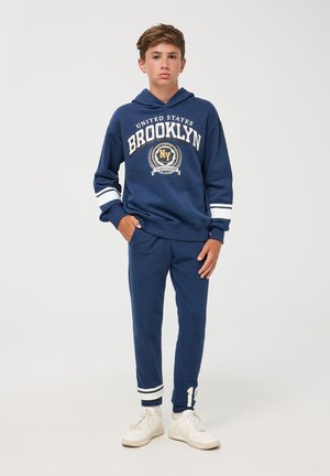 Felpa blu navy con grafica "Brooklyn", strisce bianche sulle maniche, abbinata a pantaloni jogger blu navy, con accenti bianchi e dettagli con logo.