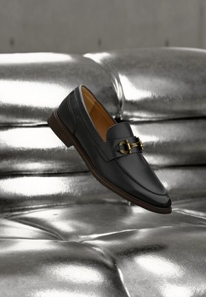 Schwarzer Lederloafer mit goldenem Schnallendetail, der über einer glänzenden, metallisch silbernen, gesteppte Oberfläche schwebt.