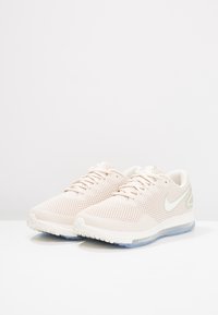 Une paire de chaussures de course Nike beige clair avec une tige en mesh, des logos swoosh blancs et une semelle translucide sur une surface blanche.