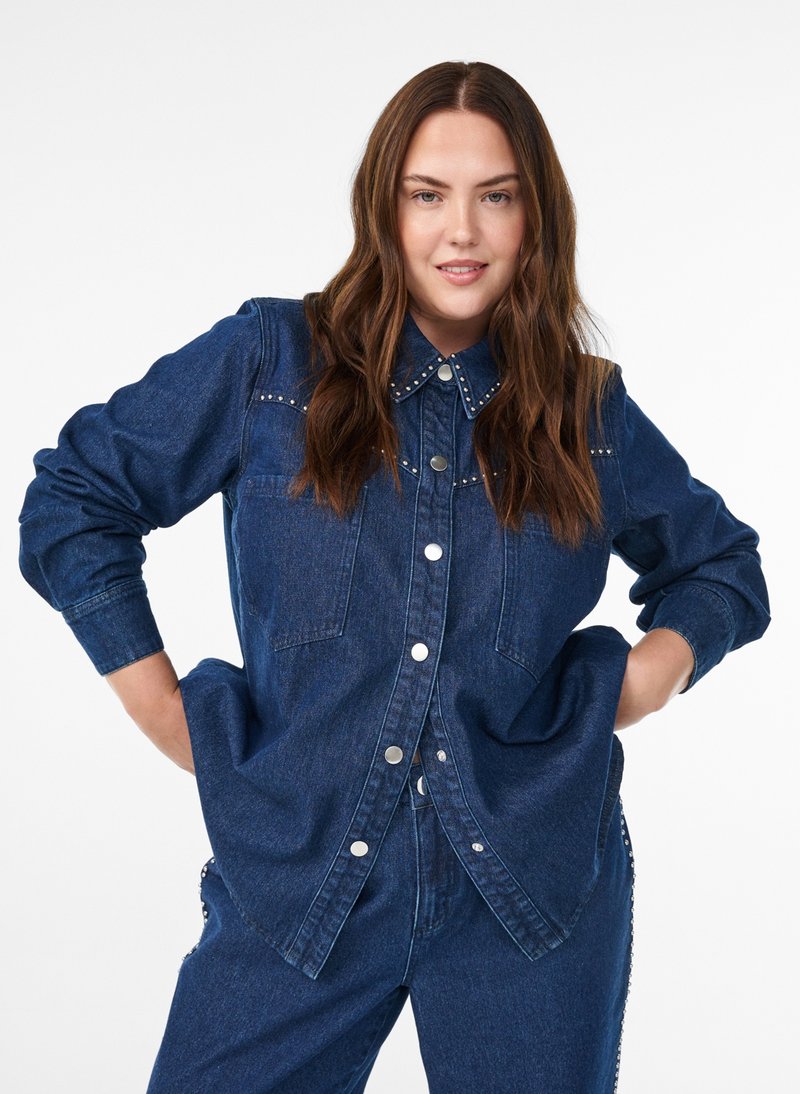 Kvinde med langt brunt hår iført mørkeblå denimskjorte og jeans med hænderne på hofterne, let smilende mod hvid baggrund.