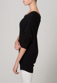 Maglione nero lavorato a maglia con design off-shoulder e scollatura embellita. Presenta maniche lunghe e una vestibilità comoda, abbinato a pantaloni bianchi.