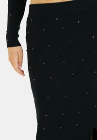 Falda negra con canalé que presenta una abertura lateral y adornada con brillantes rhinestones dispersos para un detalle adicional. Tejido suave y texturizado.