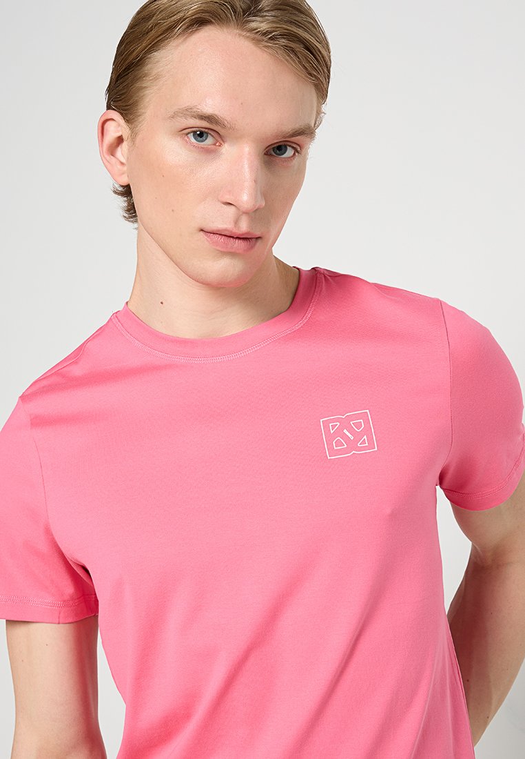 Różowa bawełniana koszulka z okrągłym dekoltem, krótkimi rękawami i małym białym geometrycznym logo na lewej piersi. Miękka tkanina.