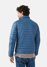 Giacca imbottita blu con collo alla coreana, design trapuntato e polsini elasticizzati; indossata sopra un maglione lavorato a maglia multicolore e jeans in denim.